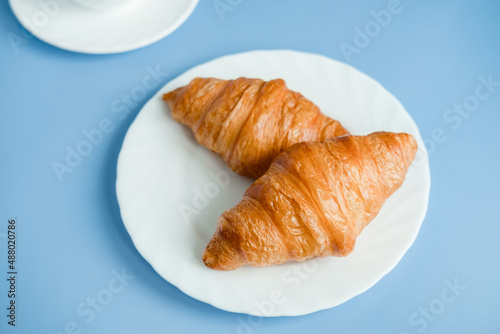 2 large croissants on a white plate, on a blue background. Morning breakfast in France. Fresh Baking with your own hands.