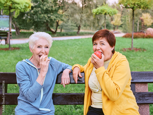 two mature women eating apples on the bench after doing sport exercises in the park. healthy lifestyle concept