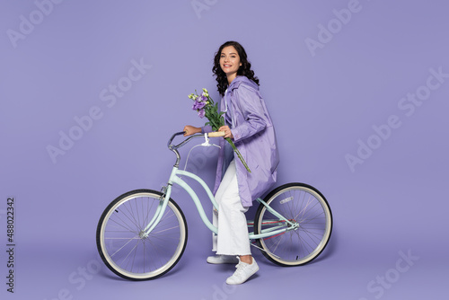 Fototapeta Naklejka Na Ścianę i Meble -  full length of cheerful young woman in violet raincoat riding bicycle and holding flowers on purple.