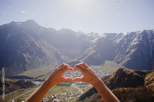 Two hands folded with their hearts against the backdrop of a mountain landscape with snow-capped peaks and a village