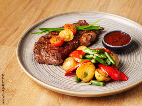 Grilled steak with vegetables and sauce. Juicy steak with tomatoes, potatoes, peppers and green beans in a plate on a wooden background. Delicious hearty lunch. Meat with baked vegetables. Close up.