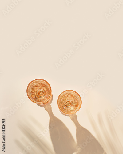 Two wine glasses from pale pink colored glass with shadows from palm leaf at sunlight, summer rest concept, white wine in trend glasses in goblets style on peach color, aesthetic top view