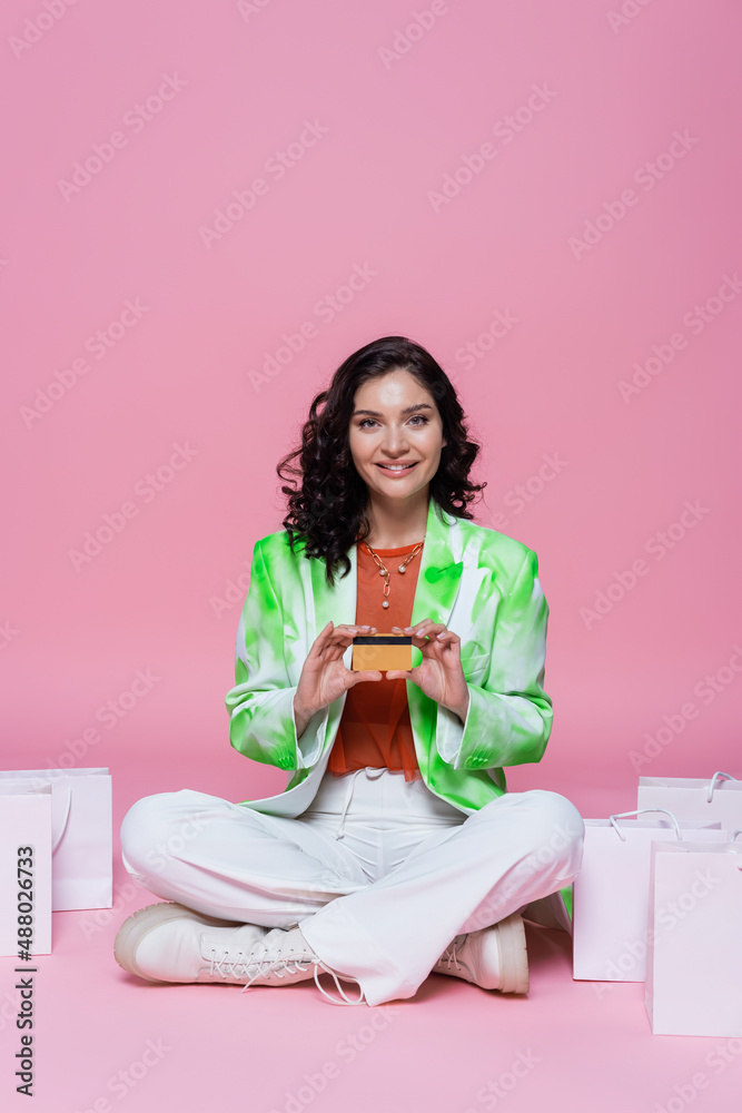 happy brunette woman in blazer sitting with crossed legs and holding credit card near shopping bags on pink. happy brunette woman in blazer sitting with crossed legs and holding credit card near shopping bags on pink.