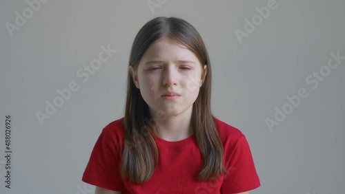 A blonde teenager girl in a red T-shirt cries and wipes her tears with her hands against the background of a white wall