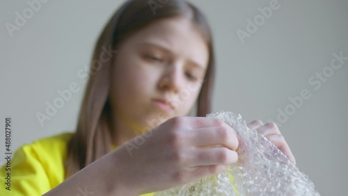 A blonde teenager girl in a yellow T-shirt bursts bubble wrap and is bored