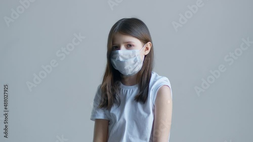 A blonde teenager girl in a white t-shirt and a blue mask touches the vaccine on her shoulder against the background of a white wall