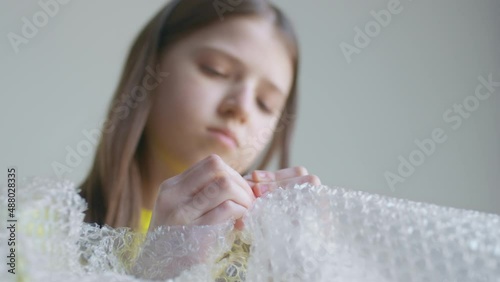 A blonde teenager girl in a yellow T-shirt bursts bubble wrap and is bored