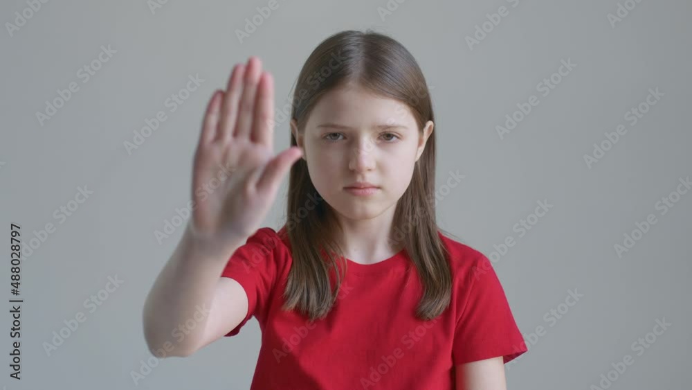 Blonde teenager girl in a red T-shirt angrily raises her hand stop in front of the background of a white wall Blonde teenager girl in a red T-shirt angrily raises her hand stop in front of the background of a white wall