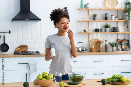 Happy african american woman standing at the cuisine table in the home kitchen drinking dietary supplements, looking away and smiling friendly, healthy lifestyle concept photo