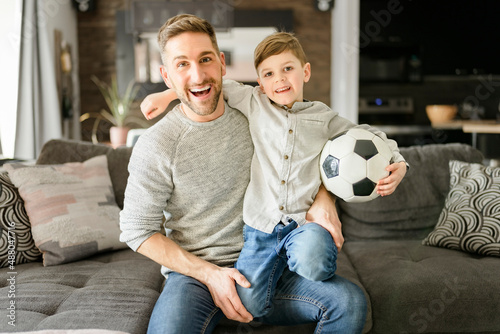 Soccer fans Emotional dad and son cheering with football ball, watching sport on tv at home