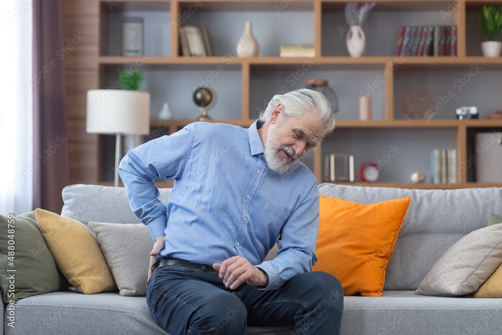 A lonely gray-haired man sits on the couch at home, has severe back pain, holds his hands on his sides and massages