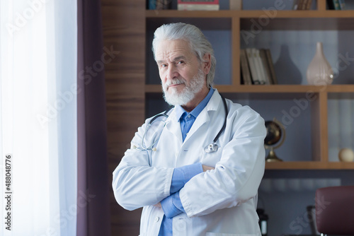 Portrait of an experienced doctor, chief gray-haired doctor of the clinic, in his office, looking at the camera with crossed arms