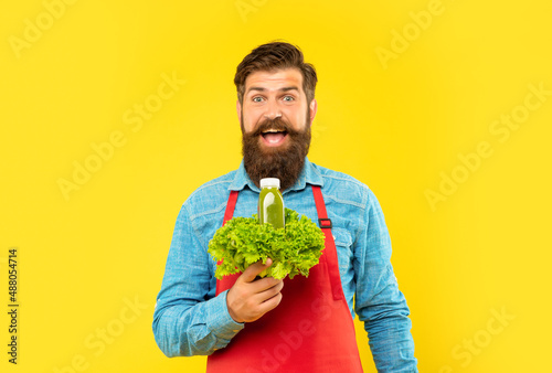 Surprised man in apron holding fresh lettuce and juice bottle yellow background, juice barkeeper