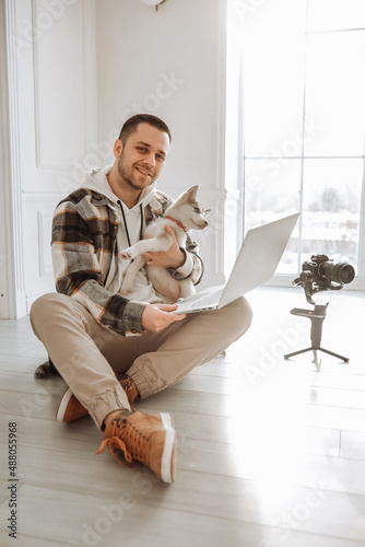 young modern videographer sitting on the floor with husky puppy near equipment with camera, stabilization system steasycam steadicam and laptop. modern european videographer style