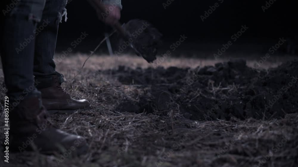 Criminal digs hole at night Young scary man digs hole in field. Close up of legs.