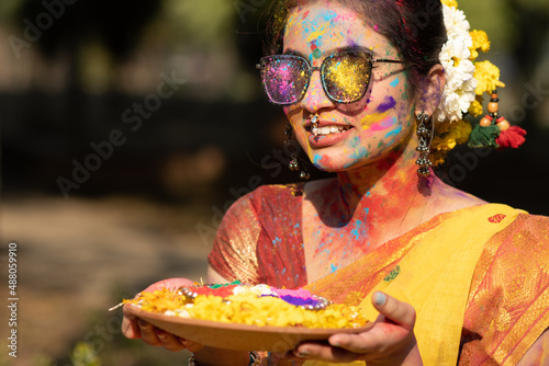 Holi festival of color, young beautiful girl or woman or lady in indian attire saree playing holi with dry organic color or colour or gulal or abeer or Holi powder placed in clay bowl with flowers