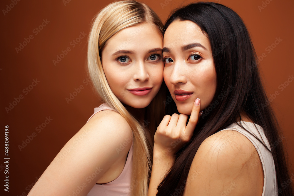 two pretty diverse girls happy posing together: blond and brunette, caucasian and asian on brown background, lifestyle people concept two pretty diverse girls happy posing together: blond and brunette, caucasian and asian on brown background, lifestyle people concept