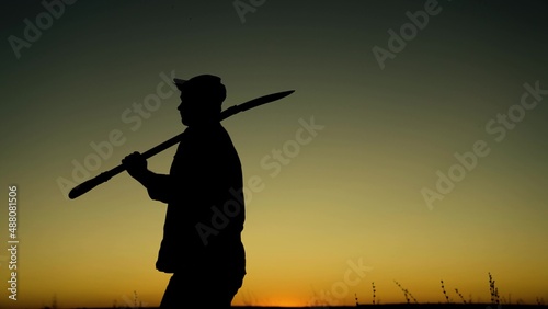 Silhouette of farmer with shovel in his hands goes through field from work. Farmer working in morning. Worker with shovel walks through plantation at sunset. Agricultural business. Growing vegetables.