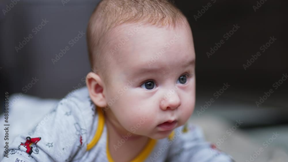 Calm peaceful baby boy lies on his belly and looks sideways. Curious toddler face portrait close up. Grey blurred backdrop.