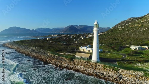 Aerial footage of a lighthouse out in a rural coastal town close to sunset. The mountains of Chapman's peak can be seen in the background. Camera slowly tracking forward and passes by the subject. photo