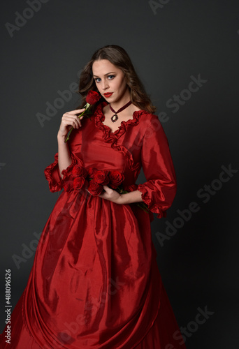   portrait of pretty female model with red hair wearing glamorous historical victorian red ballgown.  Posing with a moody dark background.