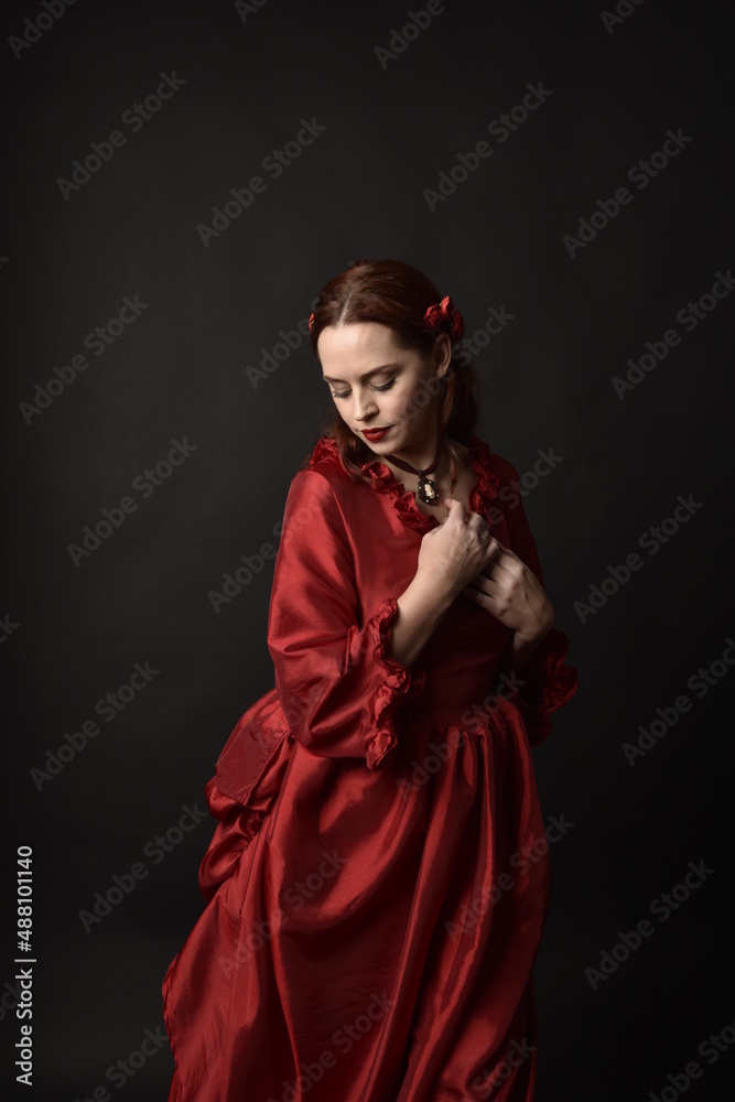   portrait of pretty female model with red hair wearing glamorous historical victorian red ballgown.  Posing with a moody dark background.