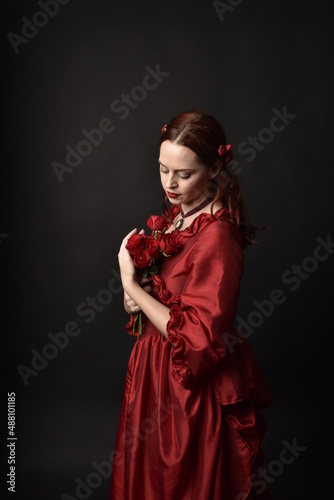  portrait of pretty female model with red hair wearing glamorous historical victorian red ballgown. Posing with a moody dark background.