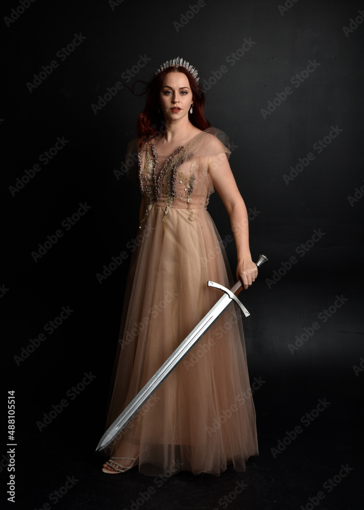Full length portrait of pretty female model with red hair wearing glamorous fantasy tulle gown and crown. Posing with a moody dark background. Full length portrait of pretty female model with red hair wearing glamorous fantasy tulle gown and crown. Posing with a moody dark background.