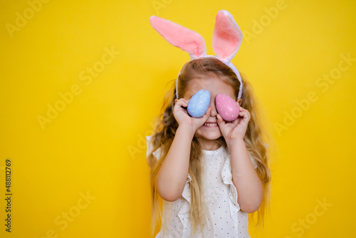 Fotografiet beautiful smiling blonde girl with bunny ears holding an easter egg in her hands, closes eyes, on a yellow background, kid celebrate easter