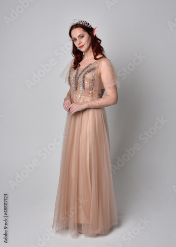  Full length portrait of pretty female model with red hair wearing glamorous fantasy tulle gown and crown. Posing with gestural arms on a studio background