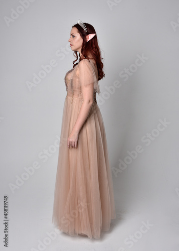 Full length portrait of pretty female model with red hair wearing glamorous fantasy tulle gown and crown. Posing with gestural arms on a studio background