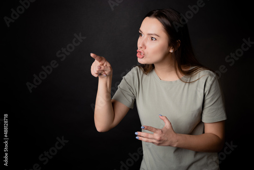 To be indignant and accuse the interlocutor, a young woman argues with the interlocutor by pointing her finger. A young brunette in a T-shirt is discussing with someone, portrait on black.