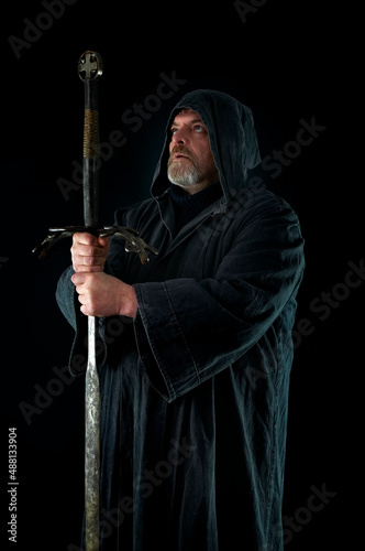 Portrait of courageous monk-warrior in black cloak with sword in hands on black background. Historical medieval templier fantasy