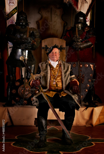 Medieval elderly count (baron, prince) sits on a throne with sword in ancient castle. Historical reconstruction in Saint Miklos castle in Ukraine