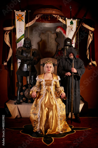 Medieval countess (baroness, princess, queen) sits on  throne in crown in  ancient castle. Behind is  monk-knight with  sword. Historical reconstruction, pretty woman in Saint Miklos castle in Ukraine