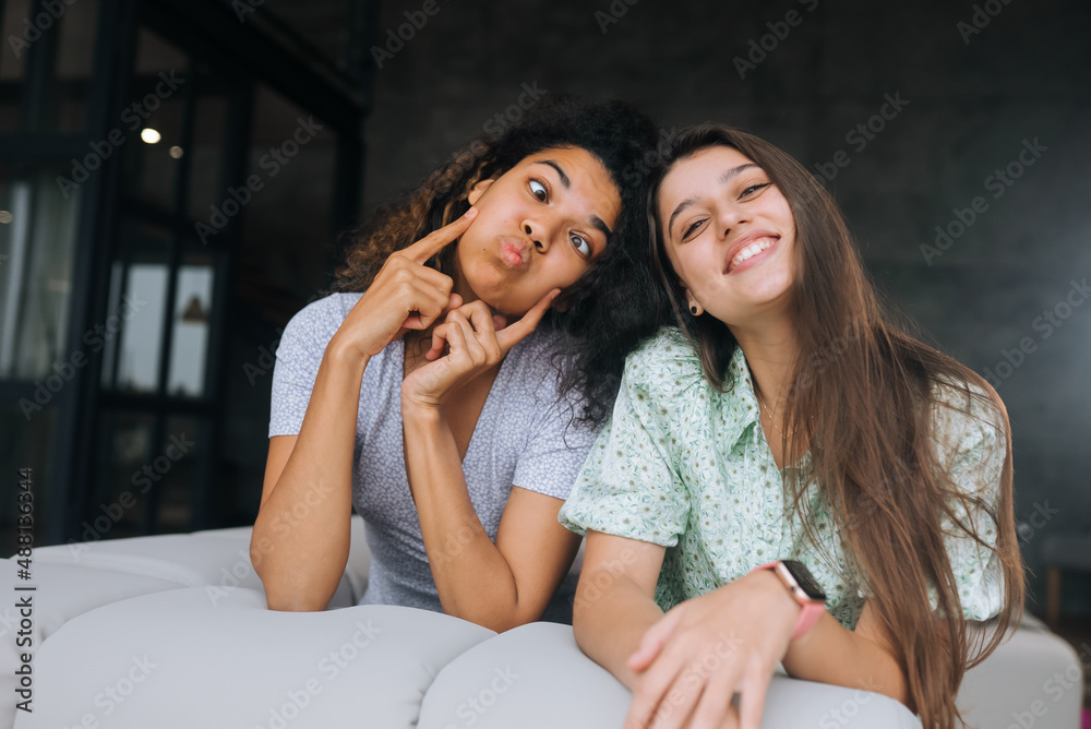 Two girls sit on the back of the sofa, look at the cameras and smile Two girls sit on the back of the sofa, look at the cameras and smile