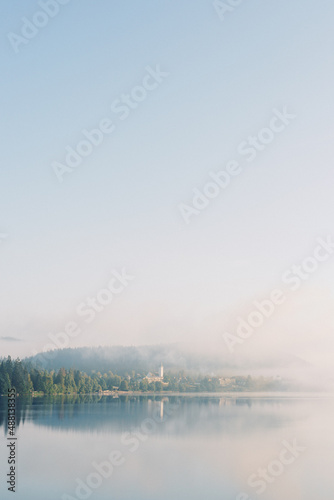 The village and lake Titisee Neustadt in Baden-Württemberg in Germany. Foggy blue lake in the morning with forest, church and town in the clouds