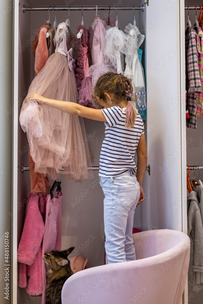 Cute little girl choosing festive dress hanged in pink cupboard