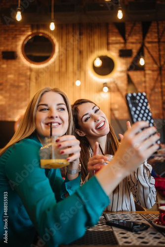 Two best friends sitting in coffee bar or restaurant after shopping and happily talking together. They are using smart phone to take selfie photo.