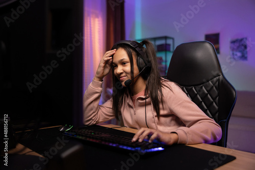 Teenager passionate about virtual world plays video shooters on pc, young girl props herself up with arm looking resignedly at monitor she has headset, backlit keyboard led purple room lights photo