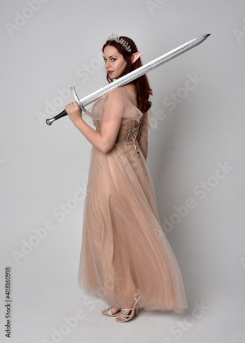  Full length portrait of pretty female model with red hair wearing glamorous fantasy tulle gown, crown. Holding a sword weapon, on studio background.