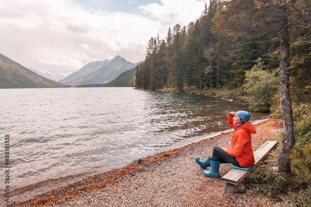 A traveler girl is sitting on a bench and enjoying a great panoramic view of a mountain lake in Altai. Hiking in the national park and solitude in nature A traveler girl is sitting on a bench and enjoying a great panoramic view of a mountain lake in Altai. Hiking in the national park and solitude in nature