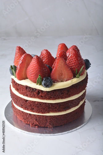 homemade fresh red velvet sponge birthday round cake with cream cheese layer and strawberry and blueberry fruits and rosemary leaf in white marble background