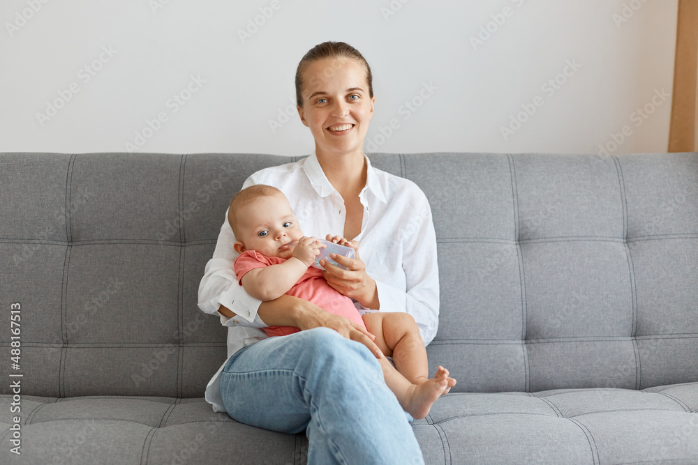 Image of smiling happy woman with bun hairstyle wearing white shirt and jeans, posing with her small daughter, looking at camera with positive facial expression. Image of smiling happy woman with bun hairstyle wearing white shirt and jeans, posing with her small daughter, looking at camera with positive facial expression.