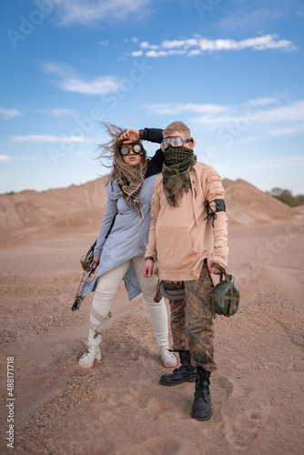 Couple in futuristic clothes in the desert. The woman has a bow, arrows and steampunk goggles, the man has a flask and a telescope. Post-apocalyptic cosplay of an archer and a nomad, artistic staged