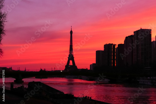 Urban landscape. View on the Eiffel tower with group of modern buildings in front of the water of Seine river. Dramatic sky with colorful clouds. Silhouette of a cityscape at sunrise. 