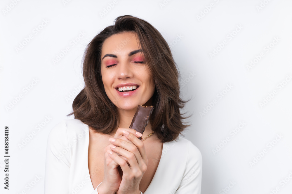 Young woman in a cocktail dress, gold chain, bright spring pink makeup on a white background. Holds a healthy cereal chocolate delicious bar. Young woman in a cocktail dress, gold chain, bright spring pink makeup on a white background. Holds a healthy cereal chocolate delicious bar.