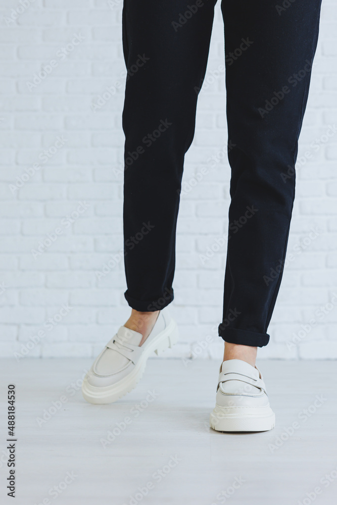 Legs of a young beautiful woman in black trousers and modern stylish white leather loafers. New collection of women's shoes spring-summer 2022 Legs of a young beautiful woman in black trousers and modern stylish white leather loafers. New collection of women's shoes spring-summer 2022