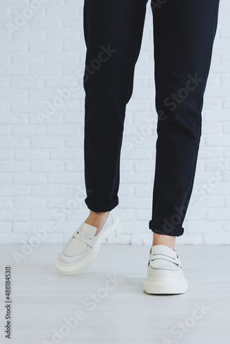 Legs of a young beautiful woman in black trousers and modern stylish white leather loafers. New collection of women's shoes spring-summer 2022