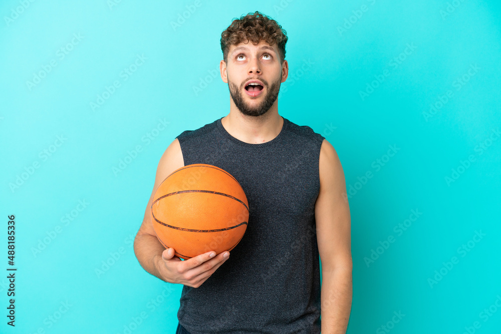 Handsome young man playing basketball isolated on blue background looking up and with surprised expression Handsome young man playing basketball isolated on blue background looking up and with surprised expression